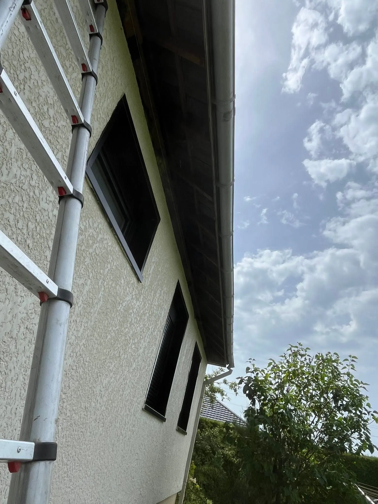 Ancienne sous-face en bois sombre et gouttière blanche le long du toit, échelle posée contre la façade