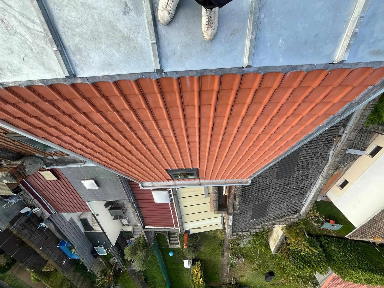 Vue plongeante sur les tuiles rouges neuves, la plateforme en zinc et les jardins voisins, avec les pieds d'un couvreur